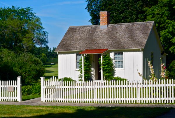 A white cottage and picket fence frames an open vista with two white grave stones and a flag in the distance.